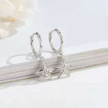 Load image into Gallery viewer, Pair of sterling silver starfish huggie earrings with cubic zirconia, displayed on a white background with a book edge.

