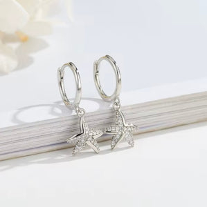 Pair of sterling silver starfish huggie earrings with cubic zirconia, displayed on a white background with a book edge.
